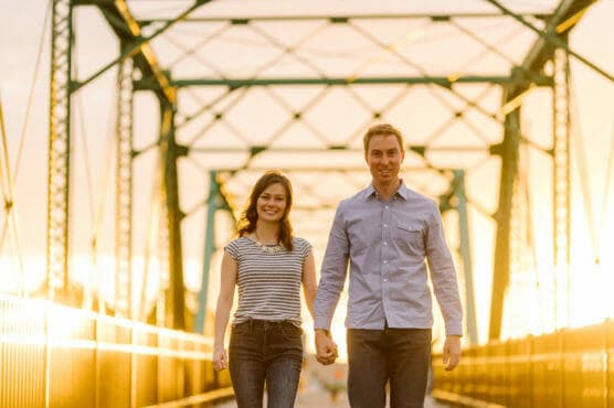 A couple walks hand in hand on a metal bridge at sunset, smiling and looking at the camera. The warm sunlight creates a golden glow around them and highlights the structure of the bridge in the background. by Justin Salem Meyer