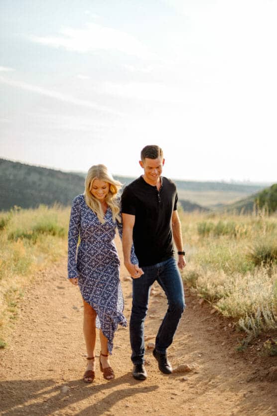 A smiling couple walks hand in hand on a dirt path through grassy hills. The woman wears a blue patterned dress and the man is in a black shirt and jeans. The sun is shining and the landscape is open and natural. by Justin Salem Meyer