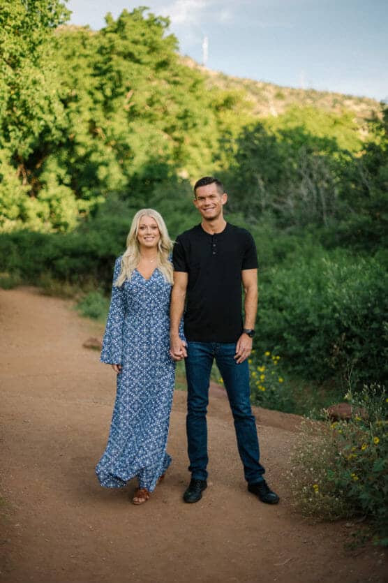 A woman in a blue patterned dress and a man in a black shirt and jeans stand holding hands on a dirt path surrounded by green trees and foliage, smiling at the camera. by Justin Salem Meyer