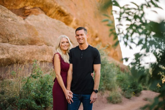 A couple stands close together on a dirt path surrounded by greenery and large reddish rock formations. The woman wears a burgundy dress and the man wears a black shirt and jeans. Both are smiling at the camera. by Justin Salem Meyer