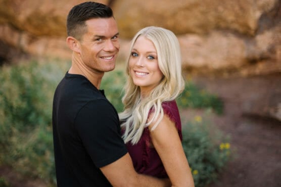 A man in a black shirt and a woman with long blonde hair in a burgundy top stand close together outdoors, smiling at the camera with greenery and rocks in the background. by Justin Salem Meyer