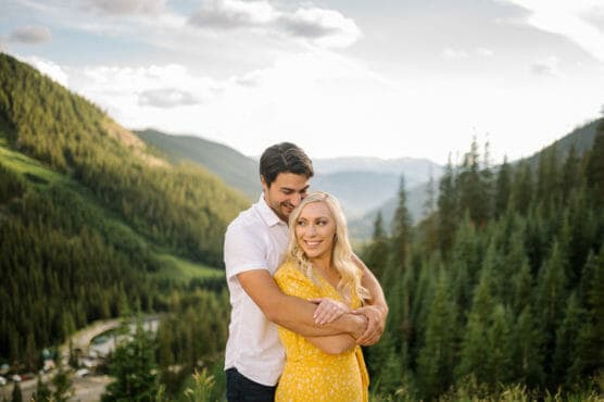 A man in a white shirt stands behind and hugs a woman in a yellow dress as they both smile, with a scenic mountain landscape and evergreen trees in the background under a bright sky. by Justin Salem Meyer