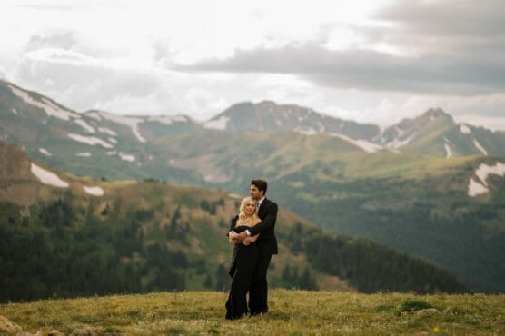 A couple dressed in formal black attire embraces on a grassy mountaintop, surrounded by scenic, green mountains and patches of snow under a cloudy sky. by Justin Salem Meyer