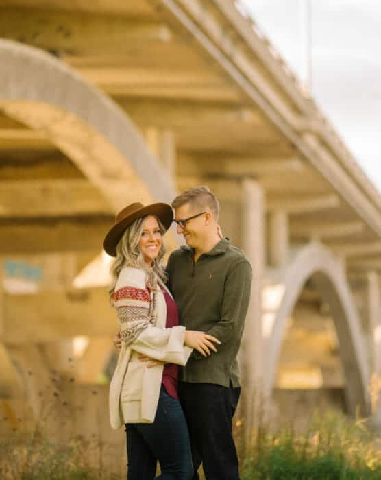 A smiling couple stands close together outdoors under a large concrete bridge. The woman wears a wide-brimmed hat, patterned sweater, and maroon shirt, while the man wears glasses and a green pullover. by Justin Salem Meyer