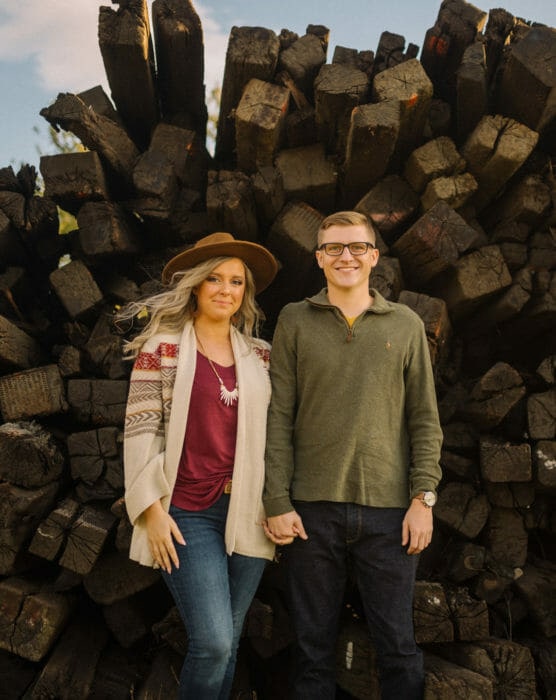 A couple stands hand in hand in front of a large stack of wooden logs. The woman wears a hat, patterned cardigan, maroon shirt, and jeans; the man wears glasses, a green shirt, and jeans. Both are smiling slightly. by Justin Salem Meyer