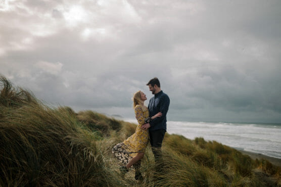 A couple stands in tall grass on a windswept coastal dune, gazing at each other and smiling, with the ocean and dramatic cloudy sky in the background. The woman wears a yellow patterned dress and the man a blue checkered shirt. by Justin Salem Meyer
