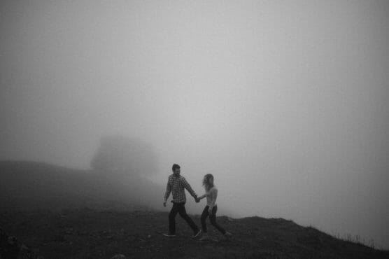 A couple holding hands walks across a grassy hilltop on a foggy day, with a tree barely visible in the background. The scene is in black and white, creating a moody and atmospheric feeling. by Justin Salem Meyer
