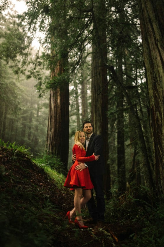 A couple stands embracing in a misty redwood forest. The woman wears a bright red dress and heels, while the man is dressed in a dark suit. Tall trees and lush greenery surround them, creating a romantic, serene atmosphere. by Justin Salem Meyer