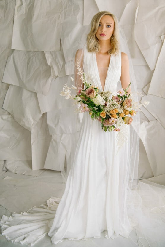 A bride in a flowing white wedding dress and veil holds a large bouquet of pastel flowers, standing in front of a textured, white paper backdrop. by Justin Salem Meyer