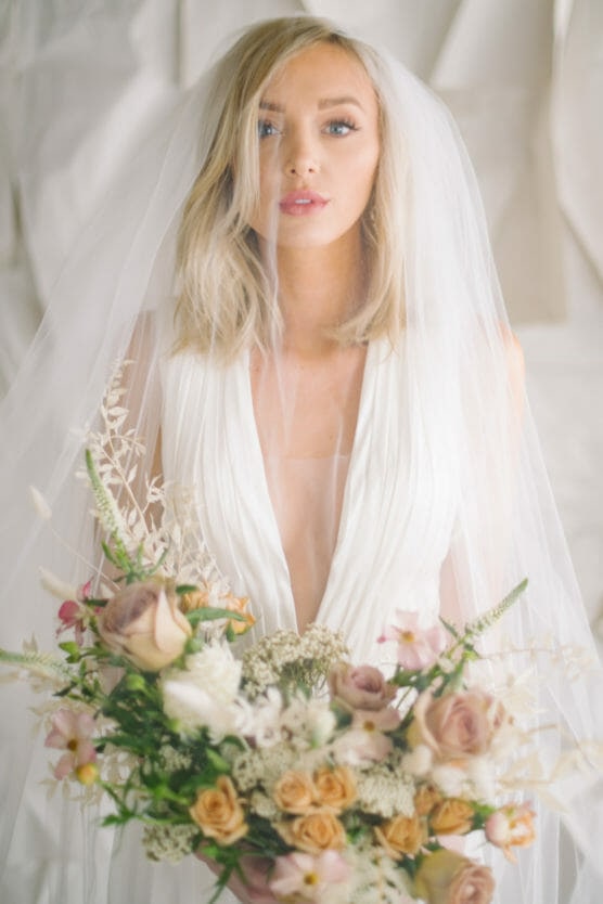 A blonde bride in a white dress and veil holds a large bouquet of pastel-colored flowers, including roses and greenery, standing against a soft, light background. by Justin Salem Meyer