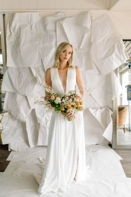 A woman in a white, sleeveless wedding dress holding a bouquet of flowers stands in front of a textured white paper backdrop, looking slightly off to the side. by Justin Salem Meyer