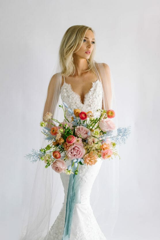 A bride in a white lace dress with a long veil holds a large bouquet of pink, peach, and orange flowers with greenery and light blue accents, standing against a plain white background. by Justin Salem Meyer