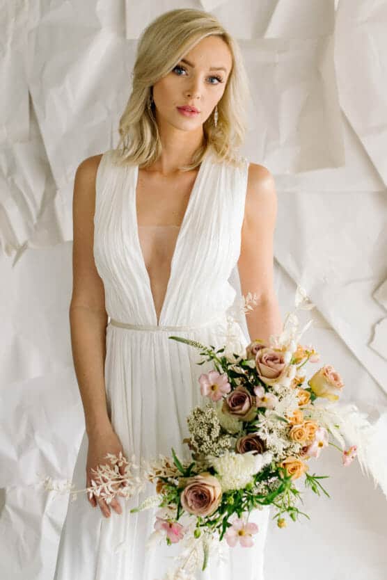 A blonde woman in a sleeveless white dress with a plunging neckline holds a large bouquet of roses and greenery, standing in front of a textured white background. by Justin Salem Meyer