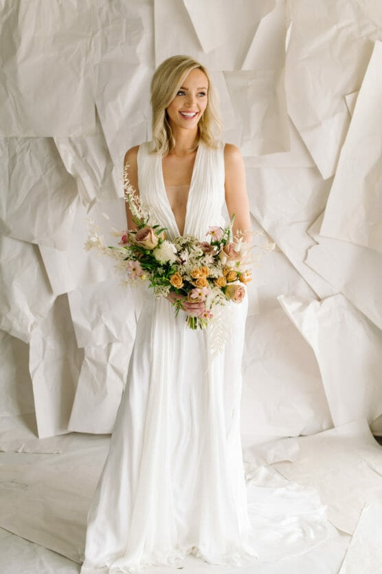 A smiling bride with blonde hair in loose waves wears a flowing white gown and holds a colorful bouquet of flowers, standing against a textured white paper backdrop. by Justin Salem Meyer