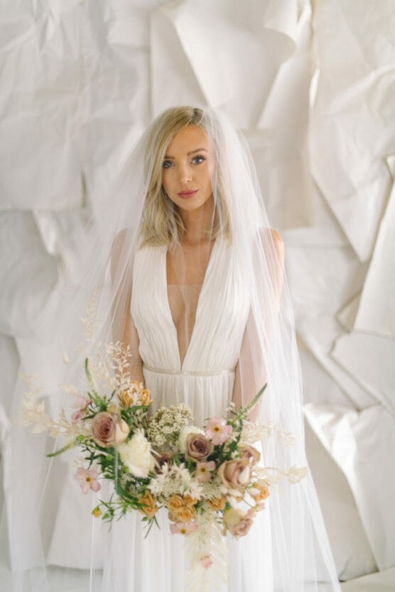 A bride with blonde hair in a white dress and veil stands holding a bouquet of pastel-colored flowers, with a white textured background behind her. by Justin Salem Meyer