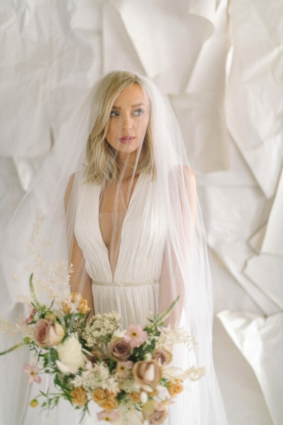 A bride with blonde hair in a white wedding dress and veil holds a bouquet of pastel and cream flowers, standing in front of a backdrop of white, textured paper. by Justin Salem Meyer