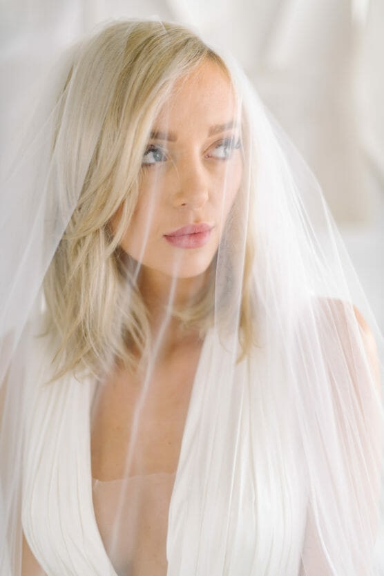 A woman with shoulder-length blonde hair wearing a white dress and a sheer veil, looking to the side with a soft expression in a bright, softly lit setting. by Justin Salem Meyer