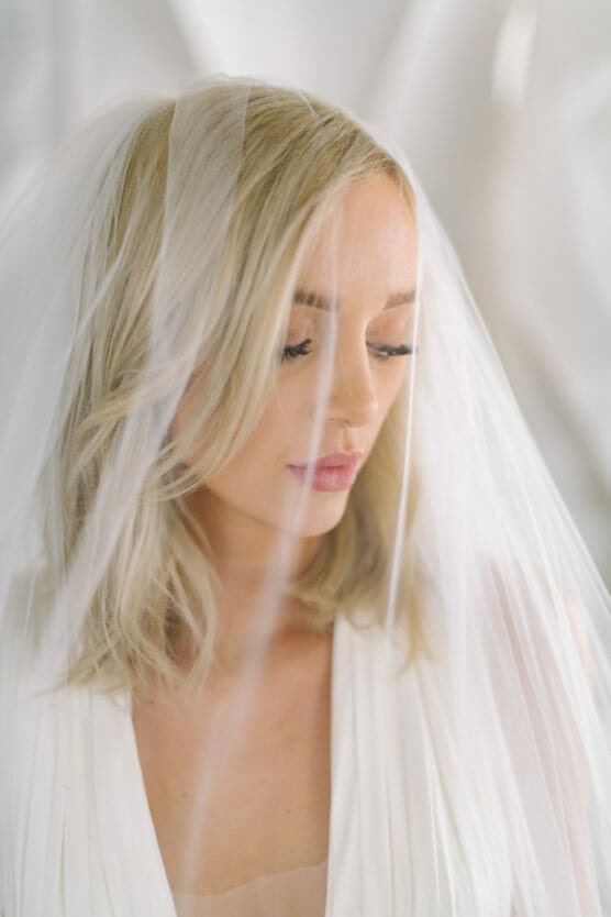 A blonde woman wearing a white bridal veil looks down softly. Her hair is loose and wavy, and she has natural makeup with pink lipstick. The background is white and softly blurred. by Justin Salem Meyer