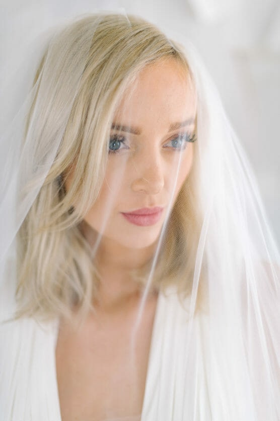 A blonde woman with blue eyes and light makeup gazes softly at the camera, wearing a white dress and a sheer bridal veil over her face. The background is bright and softly blurred. by Justin Salem Meyer
