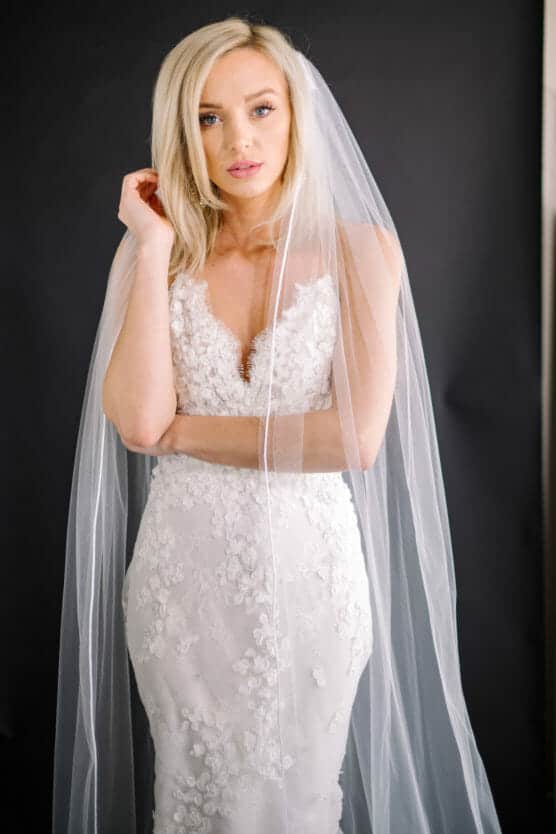 A woman in a white lace wedding dress with floral details poses against a dark background. She has blonde hair and wears a long, sheer veil that drapes over her shoulders and arms. She looks at the camera with a soft expression. by Justin Salem Meyer