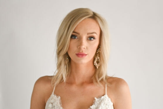 A woman with shoulder-length blonde hair, wearing statement earrings and a white, textured top, stands against a plain light gray background, looking directly at the camera with a neutral expression. by Justin Salem Meyer