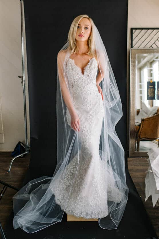 A blonde woman in a lace, fitted white wedding dress and long veil stands against a black backdrop, looking at the camera with her hand on her hip. Theres a mirror and furniture in the background. by Justin Salem Meyer