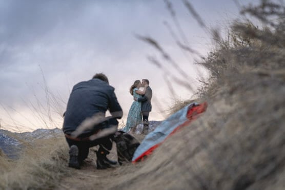 A photographer crouches in tall dry grass, capturing a couple sharing a kiss on a hillside at sunset. A colorful blanket and backpack lie on the ground nearby. by Justin Salem Meyer