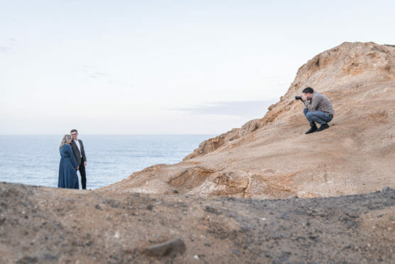 A photographer crouches on a rocky slope, taking photos of a couple standing together near the ocean under a pale sky. by Justin Salem Meyer