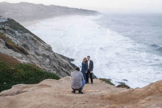 A photographer crouches on a rocky cliff to take a picture of a smiling couple standing near the edge, with waves crashing on the shoreline and hills in the background. by Justin Salem Meyer