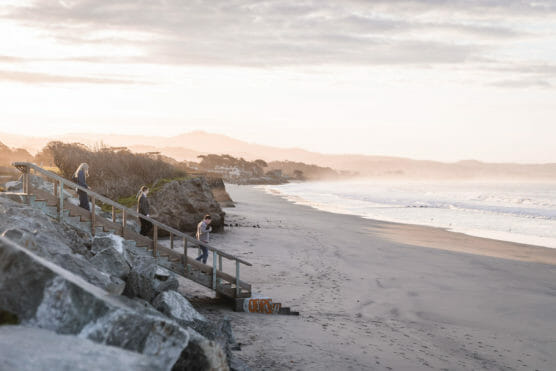 A few people walk down wooden stairs to a quiet, sandy beach at sunrise or sunset. Gentle waves wash ashore, and distant mountains are visible under a cloudy sky. by Justin Salem Meyer