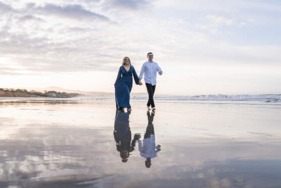 A couple holds hands while walking barefoot along a wet sandy beach at sunset, with their reflections visible on the shore and waves in the background. by Justin Salem Meyer