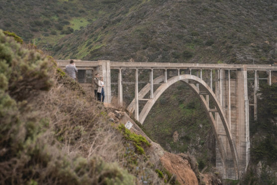 Two people stand on a rocky ledge taking photos near a large concrete bridge that spans a deep, green canyon, with mountains in the background. by Justin Salem Meyer