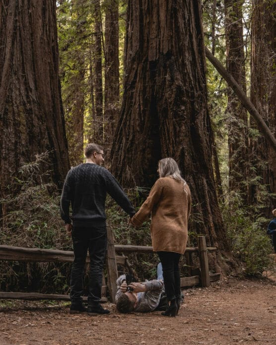 A couple holding hands stands on a forest trail, while another person lies on the ground taking their photo. Towering redwood trees surround them. by Justin Salem Meyer
