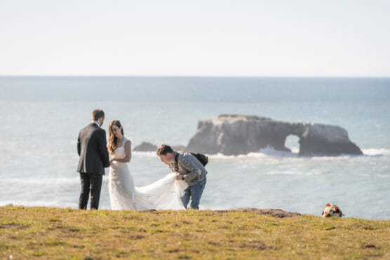 A bride and groom stand on a grassy cliff overlooking the ocean, while a person adjusts the bride’s dress. A rock formation with an arch is visible in the water, and a bouquet lies on the grass nearby. by Justin Salem Meyer