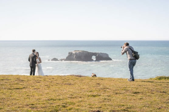 A photographer takes pictures of a bride and groom standing together on a grassy cliff, overlooking the ocean with a large rock formation in the water in the background. by Justin Salem Meyer