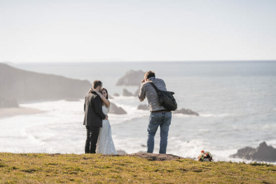 A photographer takes pictures of a couple in wedding attire standing on a grassy cliff overlooking the ocean, with a bouquet of flowers on the ground nearby. by Justin Salem Meyer