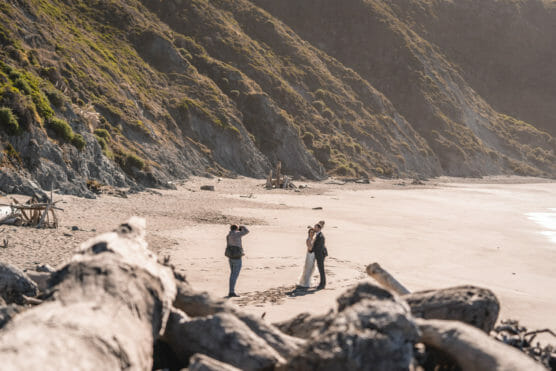 A photographer captures a couple embracing on a sandy beach with rocky cliffs and driftwood in the background, under natural sunlight. by Justin Salem Meyer
