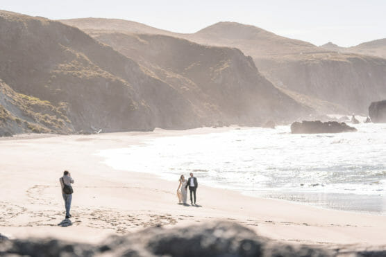 A couple in wedding attire walks hand in hand along a sandy beach, with cliffs in the background and waves rolling in. A photographer stands nearby, capturing the moment on a bright, sunny day. by Justin Salem Meyer