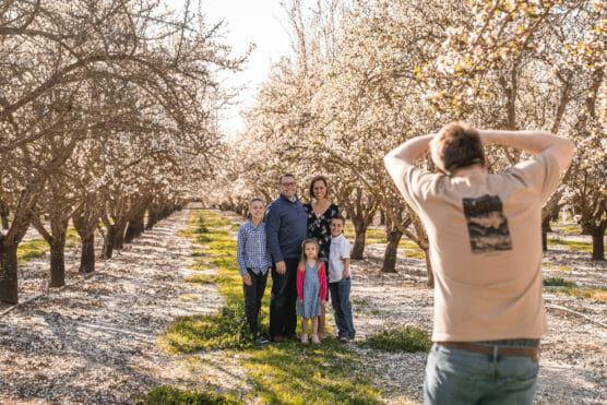 A photographer takes a picture of a family of five—two adults and three children—posing together in a blooming orchard with rows of flowering trees. The sun is shining, casting a warm light over the scene. by Justin Salem Meyer