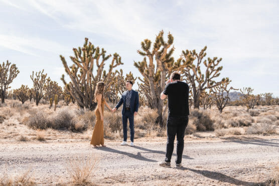 A photographer takes pictures of a couple standing on a dirt road in a desert landscape with Joshua trees and dry bushes under a bright, clear sky. by Justin Salem Meyer