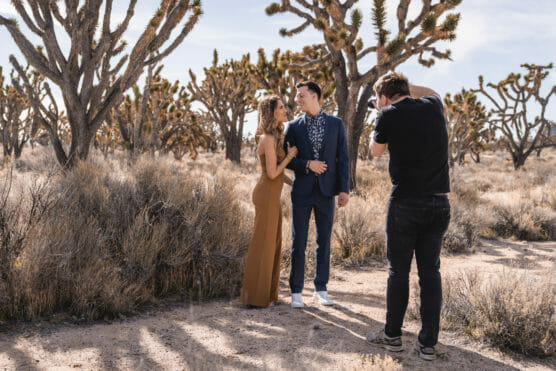 A photographer takes pictures of a couple posing in a desert landscape with Joshua trees. The woman wears a long brown dress, and the man wears a blue suit. Its sunny with dry bushes around them. by Justin Salem Meyer