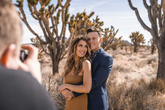 A smiling couple poses in a desert landscape with Joshua trees, as a photographer takes their picture. The woman wears a tan dress and the man wears a navy suit, standing close and embracing. by Justin Salem Meyer