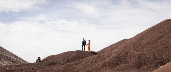 Two people stand together on a rocky, brown hill under a cloudy sky, holding hands; another person sits lower down on a nearby mound, facing them. The landscape appears vast and barren. by Justin Salem Meyer