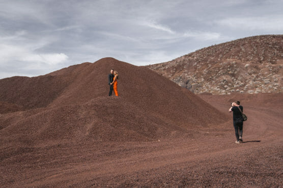 A photographer takes a picture of two people standing on a large mound of reddish-brown gravel, with a barren, rocky hillside and a cloudy sky in the background. by Justin Salem Meyer