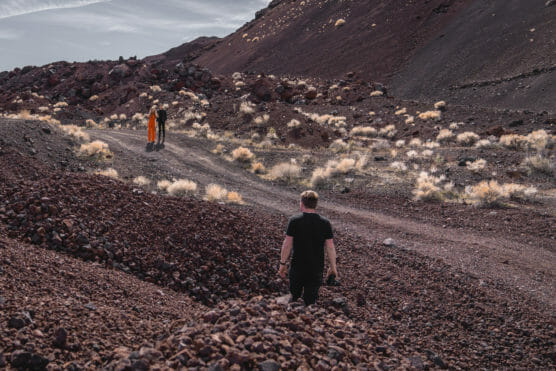 A person in black walks along a rocky, reddish-brown desert path toward two people in orange and black standing in the distance, surrounded by sparse dry bushes and rugged hills. by Justin Salem Meyer