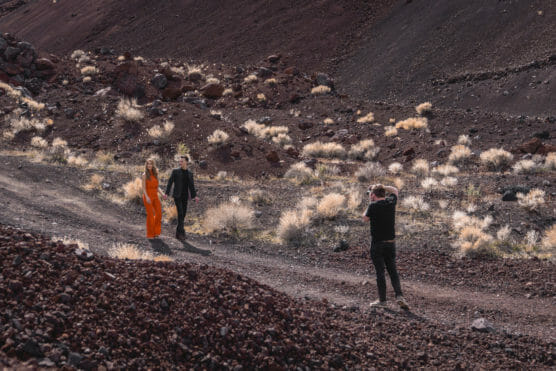 A photographer takes pictures of a woman in an orange dress and a man in a dark suit as they walk on a rocky, desert-like landscape with scattered dry bushes. by Justin Salem Meyer