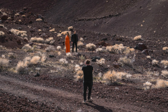 A photographer takes a picture of a couple standing on rocky, sparsely vegetated terrain. The woman wears a bright orange dress, and the man is dressed in black. Dry grass and reddish-brown earth surround them. by Justin Salem Meyer