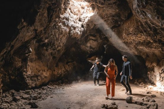 Three people walk inside a large, rocky cave with sunlight streaming through an opening above, illuminating the cave floor and walls. by Justin Salem Meyer