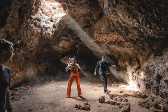 A woman in orange pants and a man in dark clothing stand inside a rocky cave, illuminated by sunlight streaming through an opening above. Another person stands partially visible on the left, watching them. by Justin Salem Meyer