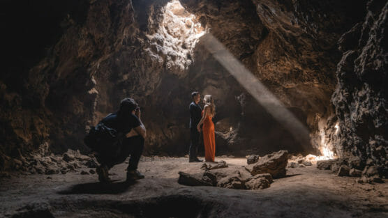 A photographer captures a couple standing close together inside a dimly lit cave, with a beam of sunlight shining down from an opening above, illuminating them. Rocks and rough cave walls surround the scene. by Justin Salem Meyer